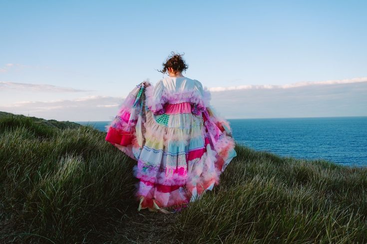 a woman stands on a grassy hill overlooking the ocean, wearing a colorful, frilly dress. she is facing away from the camera. the dress has puffy sleeves and ruffles. the sky is light blue with a few clouds. the landscape consists of grass and the ocean. the horizon is visible in the background.