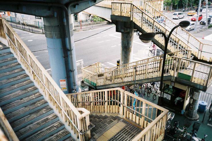 the image shows a highangle view of a pedestrian overpass in an urban japanese setting. the overpass features wooden and metal railings, stairs leading up and down, and several concrete support pillars.  numerous bicycles are parked underneath the overpass.  in the background, a busy street with cars is visible. the overall scene is a typical example of urban infrastructure in a dense city. the lighting suggests it is daytime.