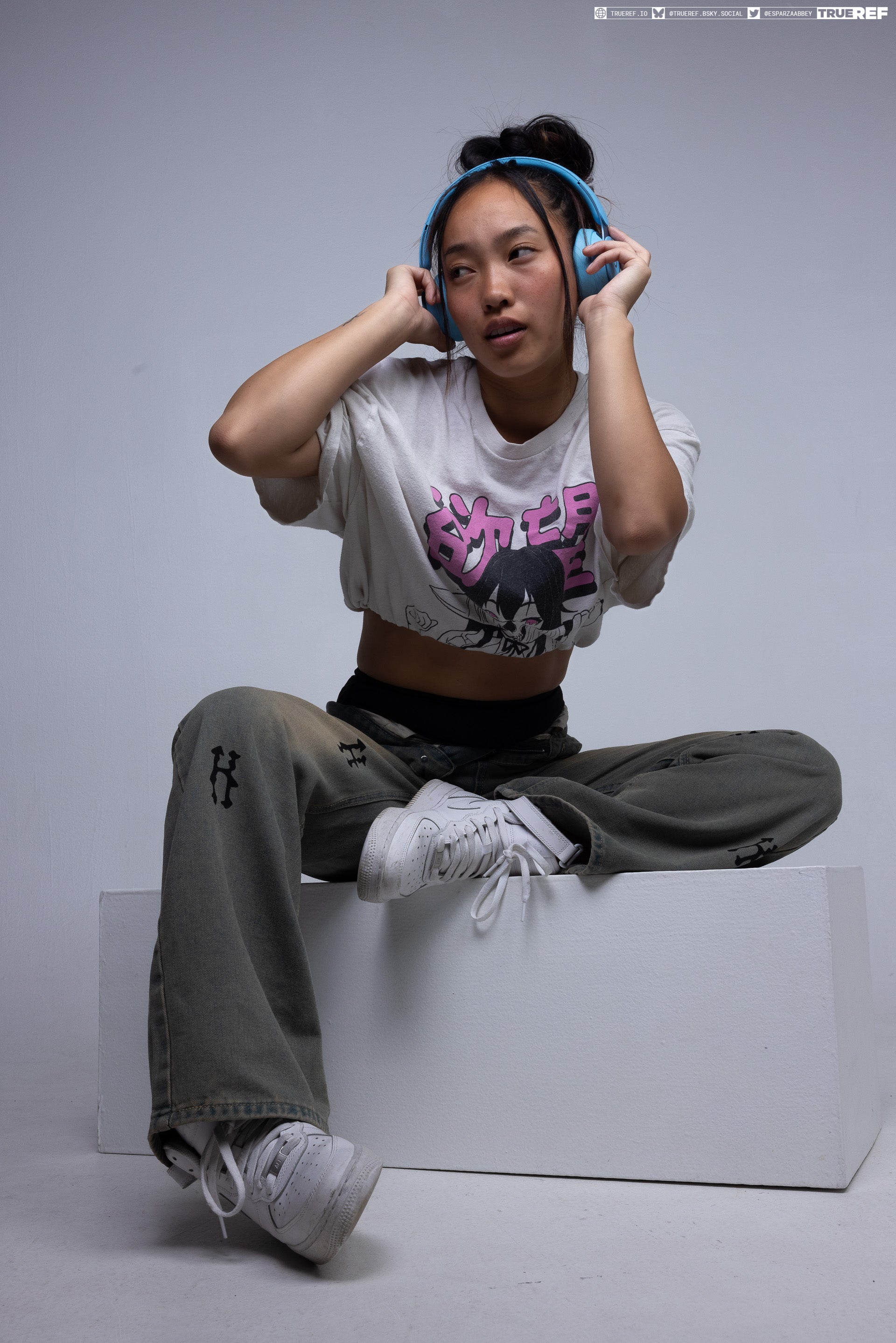 a young asian woman is sitting crosslegged on a white cube in a studio setting. she's wearing light blue headphones, a white graphic crop top, and baggy olivegreen jeans with designs on the legs. her footwear consists of white sneakers. the background is a plain grey.