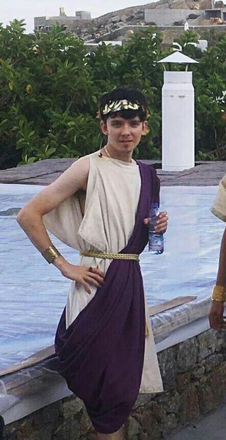 a young man dressed in a roman toga, complete with a laurel wreath, holding a water bottle. he appears to be outdoors in a scenic location.