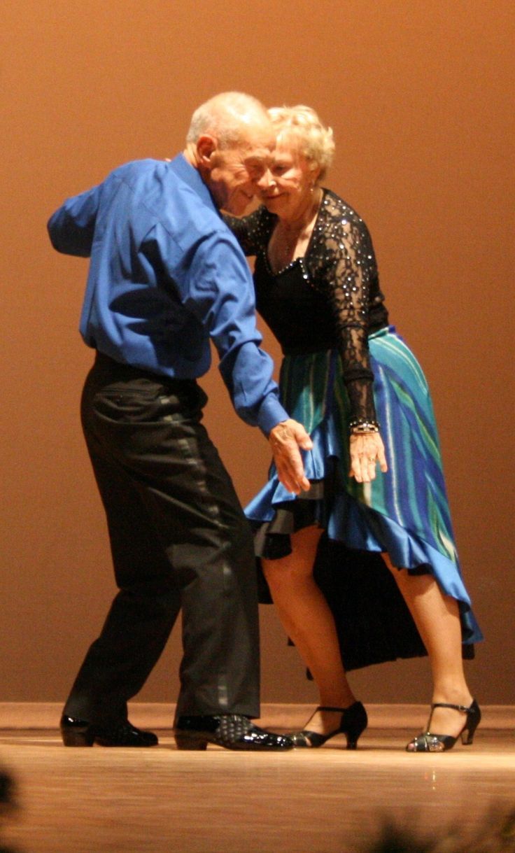 a man and woman in formal attire are dancing together on stage. the man is wearing a blue shirt, black pants, and black shoes. the woman is wearing a black top with a blue and white dress and black shoes. they are smiling and looking happy together. the man is holding the woman's hand as they dance.