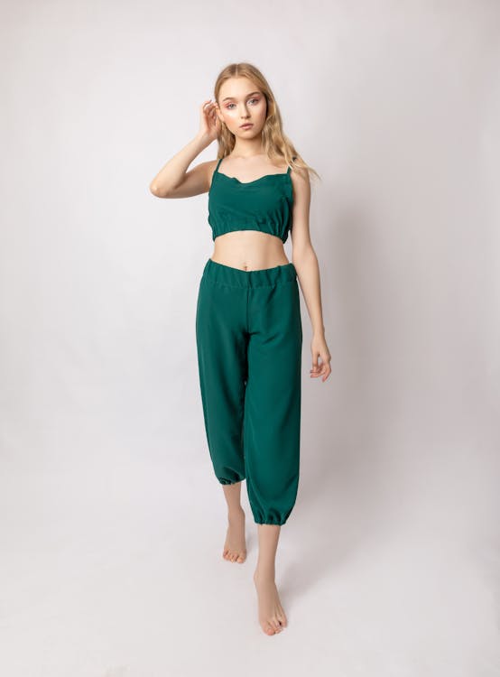 a fulllength studio shot of a young blonde woman model posing against a white background. she is wearing a stylish emerald green twopiece outfit, consisting of a cropped top and loosefitting, highwaisted pants. the clothing appears to be made of a soft, flowing fabric. the model stands with her body slightly angled, one hand gently touching her hair, looking directly at the camera. her expression is calm and natural. the overall feel of the image is elegant yet casual.