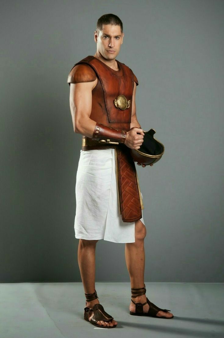 a man stands in a studio, dressed in an ancientstyle costume. he wears brown leather armor, a white skirt, and sandals. he holds a black pottery bowl in his hands. the background is a plain gray.