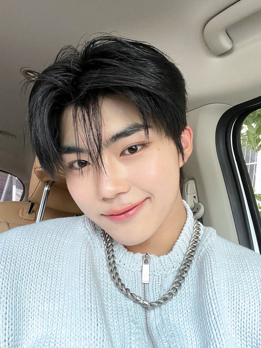 a closeup selfie of a young asian man with a trendy hairstyle and a silver chain. he is wearing a light blue sweater and is smiling slightly at the camera. the background is the interior of a car, and the lighting is soft and flattering.