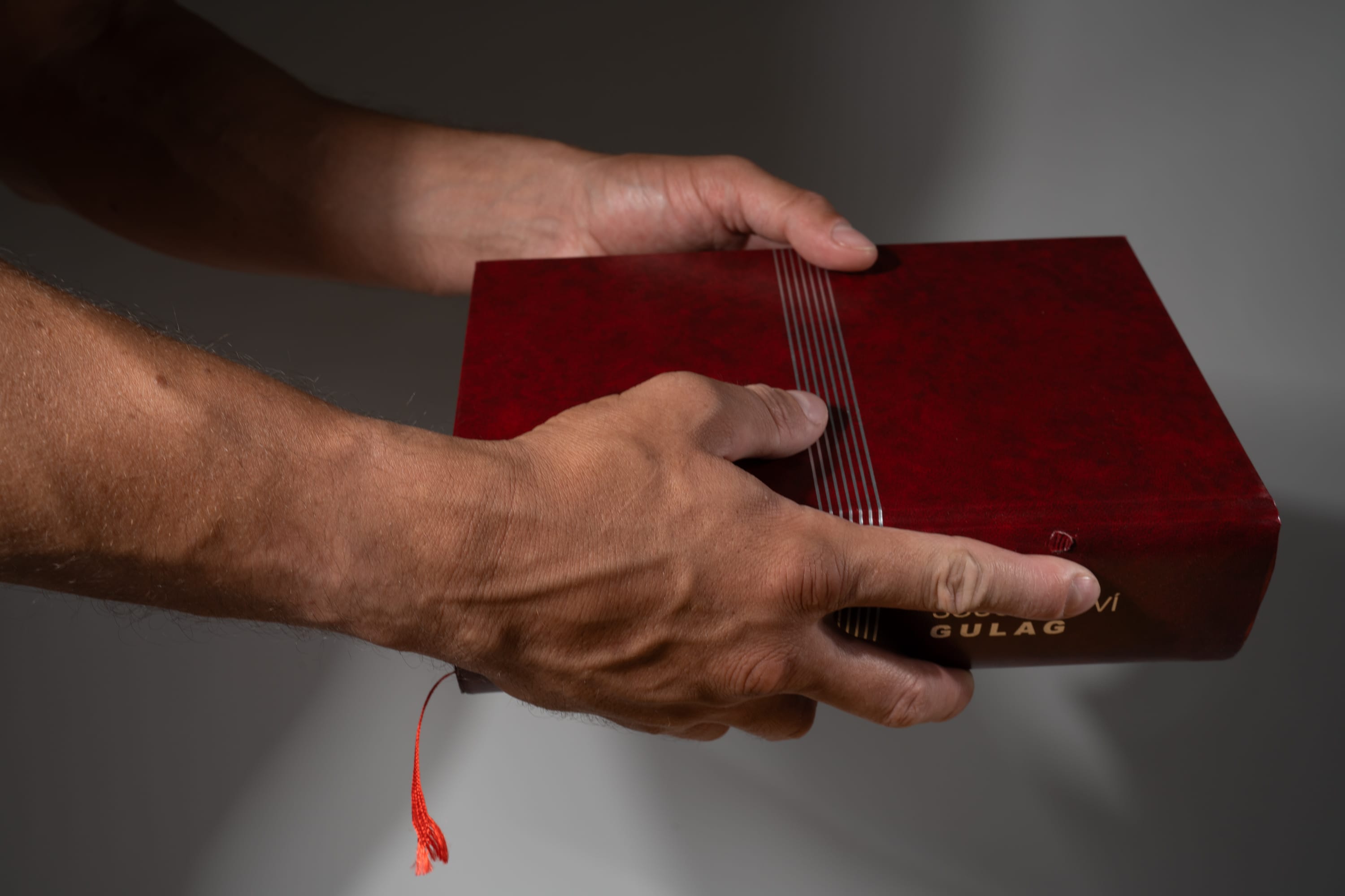 a man holds a red book titled "gulag" with both hands. the book appears to be old and worn, with a leather cover and gold lettering. the man's hands are strong and weathered, suggesting age and experience. the background is a neutral gray, drawing attention to the book and the hands holding it.