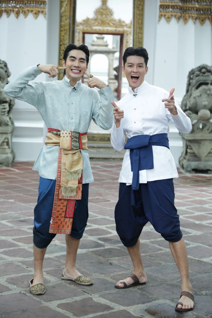 two men wearing traditional thai clothing stand in front of a temple, smiling for the camera,  while sightseeing in southeast asia.