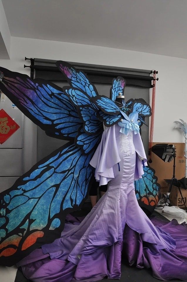 a dress with butterfly wings is displayed on a mannequin. the dress is a long, formal gown with a lavender color scheme, transitioning from light to dark hues. attached to the dress are large, ornate butterfly wings, predominantly blue with black outlines and accents of purple, orange, and green. the mannequin is positioned in what appears to be a studio setting with various background elements including a black backdrop, equipment, and storage boxes.