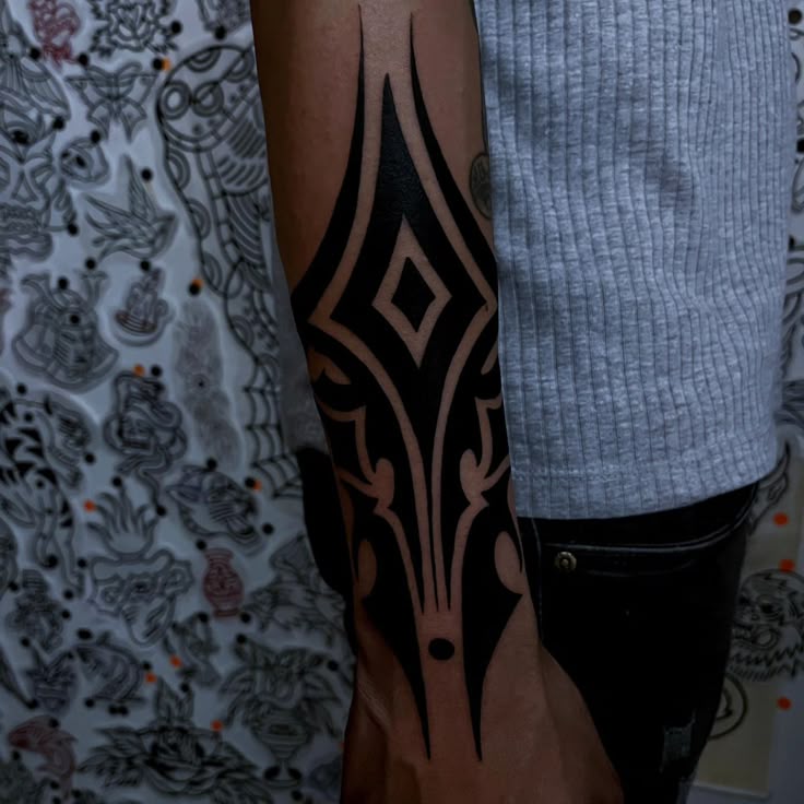 the image shows a person's arm with a tribalstyle black tattoo on their forearm. the tattoo features a diamond shape in the center, surrounded by flowing lines and patterns. the person is wearing a gray shirt and black pants. the background has a patterned wallpaper.
