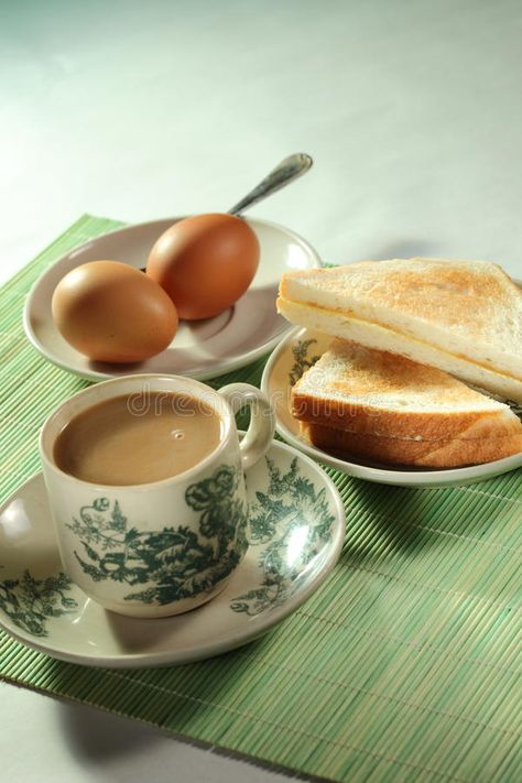a breakfast scene with a cup of tea or coffee, two soft boiled eggs, buttered toast and a green place mat on a white table.  a silver spoon rests on the plate the eggs are on.