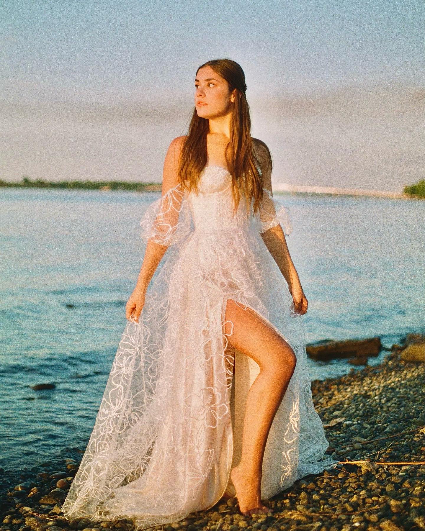 a young woman in a beautiful offtheshoulder, lace wedding dress stands on a pebble beach at sunset.  the dress is long and flowing, with a thighhigh slit. the woman is barefoot and looks out toward the water. the overall mood is romantic and serene. the golden hour light enhances the beauty of the scene and the dress.
