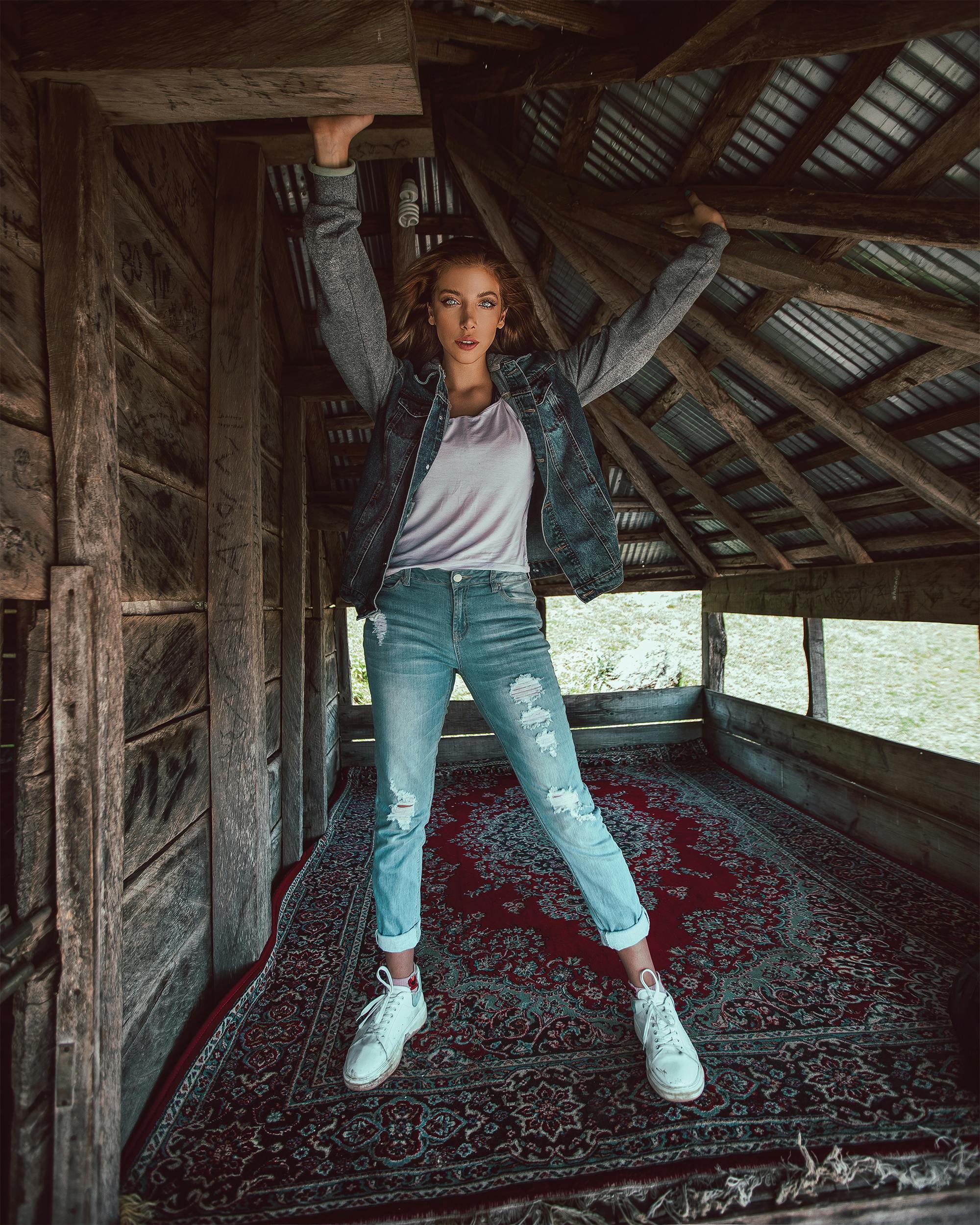 a fullbody shot of a young woman posing inside a rustic wooden structure. she's wearing a denim jacket, lightwash ripped jeans, and white sneakers. her arms are raised above her head, holding onto the wooden beams of the structure. the scene is set on a red and darkcolored patterned rug, with the background highlighting the aged wood and metal roofing of the building.  the overall style is casual and fashionoriented.