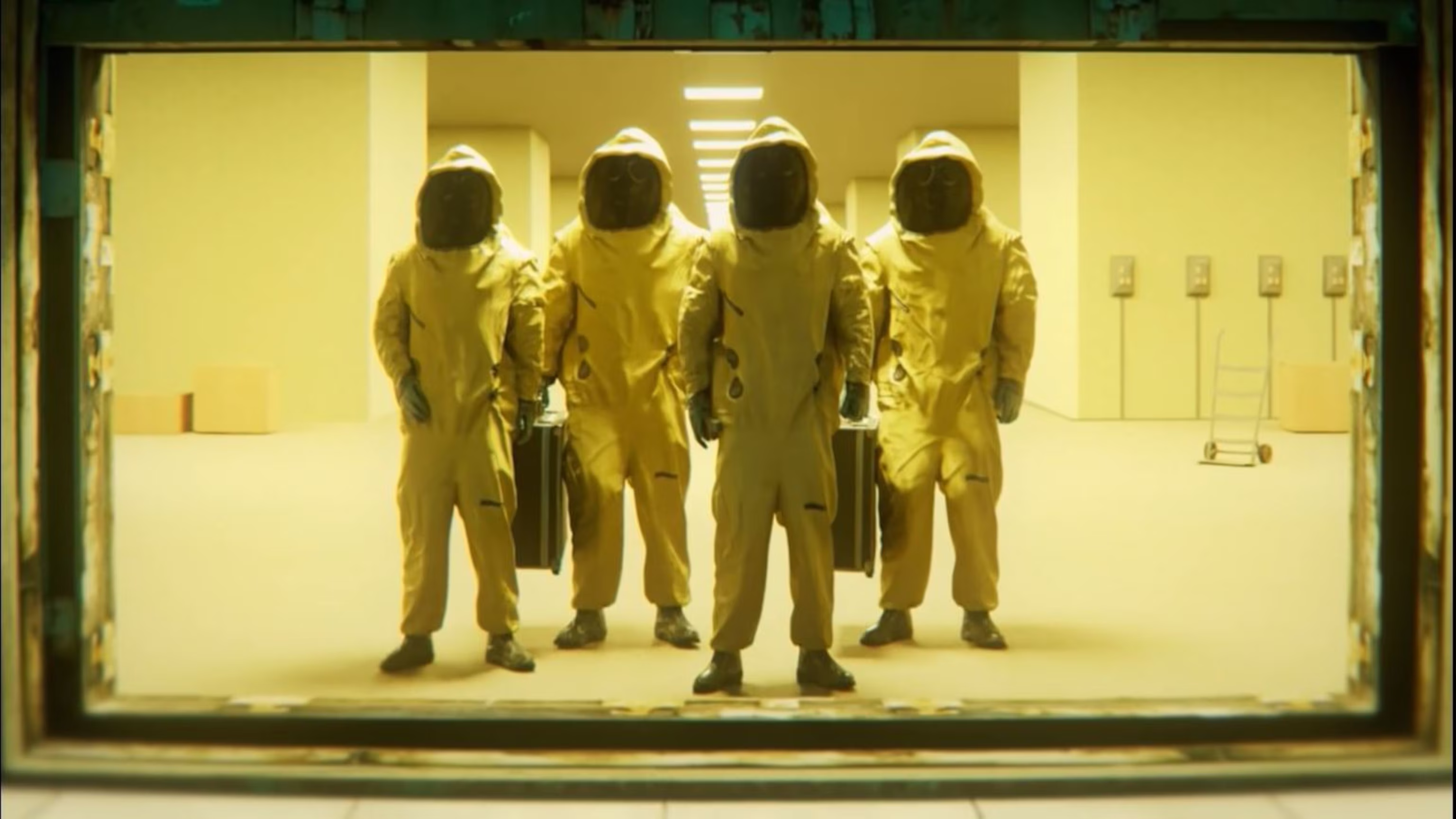 a view from a doorway into a sterile, brightly lit, white hallway.  there are five people standing in the hallway, all wearing yellow hazmat suits, facing forward. the people are standing in a row, with the person in the middle positioned closest to the camera. the hallway is empty, except for a small hand truck leaning against the wall on the right side of the frame.  there is a large, flat, white box directly behind the hand truck.  a doorway is visible at the end of the hallway.  the person on the far left is slightly out of focus, while the remaining people are clear and welllit.