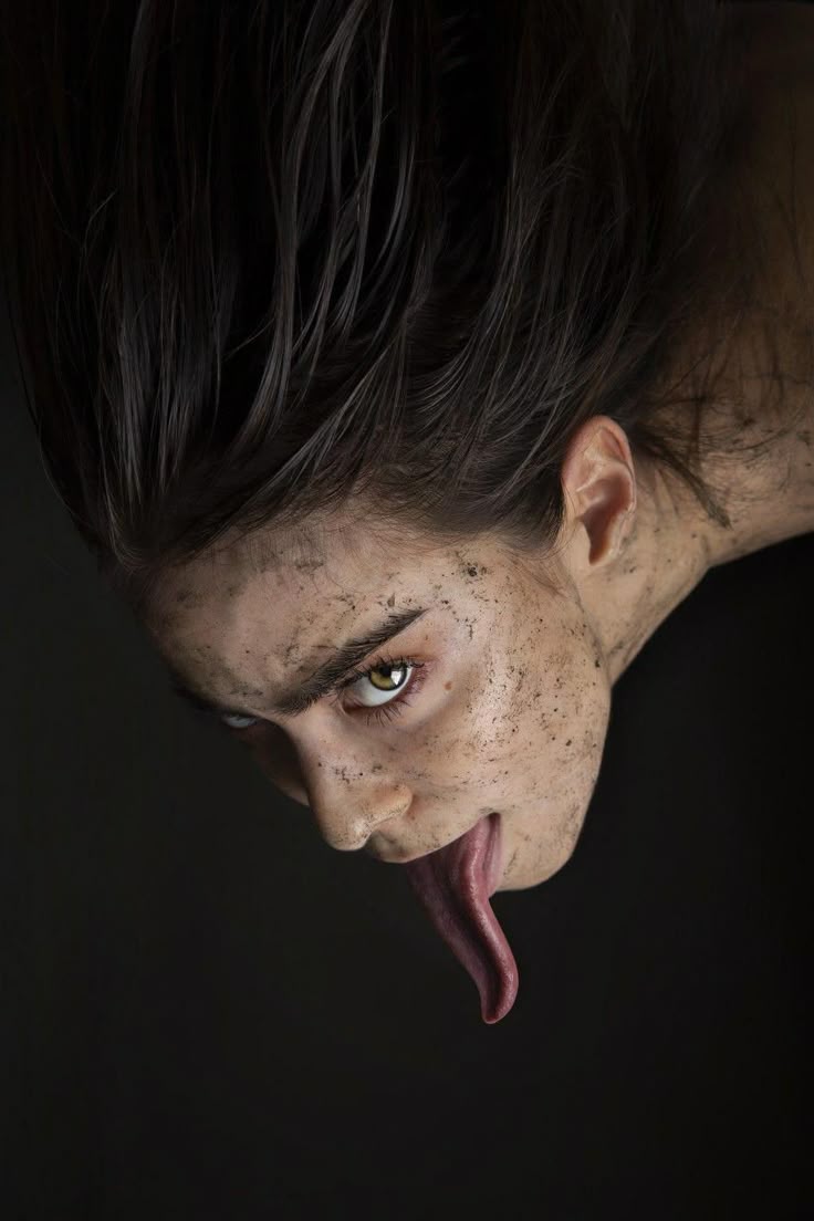 a portrait of a woman with long dark hair looking down with an elongated tongue. her face is covered in dirt or flecks, and her expression appears unsettling. the background is black, creating a stark contrast.