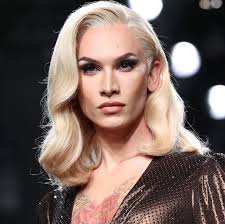 loseup portrait of a transgender fashion model with shoulderlength blonde wavy hair styled in a glamorous fashion. she is wearing a dark brown, sparkly top and has striking makeup. her expression is serious and confident as she poses for the camera. the background is blurry and dark, typical of a fashion show or photoshoot setting. the focus is on the model's face and upper body.