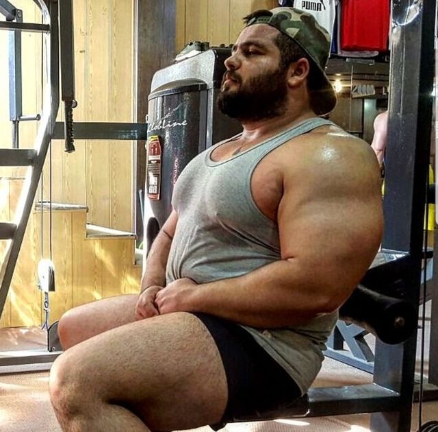 a muscular man is sitting on a weightlifting machine in a gym. he is wearing a gray tank top and black shorts. he has a beard and a camo hat on. he looks to be taking a break after a workout.