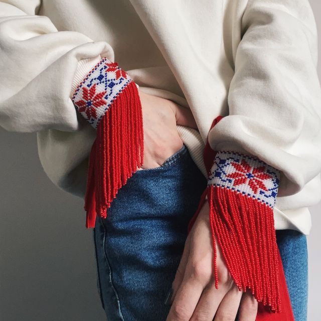 a closeup shot of a woman's hands wearing red and white beaded bracelets with floral embroidery on each wrist. the bracelets have tassels. the woman is wearing a white sweater and blue jeans. the image is captured in a casual and closeup manner, highlighting the details of the bracelets and the woman's hands. the image evokes a bohemian and folk style, with the colorful beaded bracelets adding a touch of vintage charm.