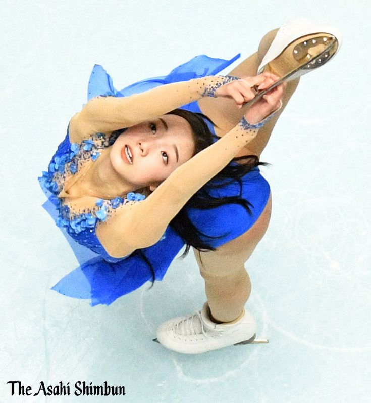 a female figure skater in a blue dress performs a spin on the ice. she is in midair, with her right leg extended and her left leg bent. her skates are white and her dress is decorated with blue flowers. she is concentrating intently on her performance.