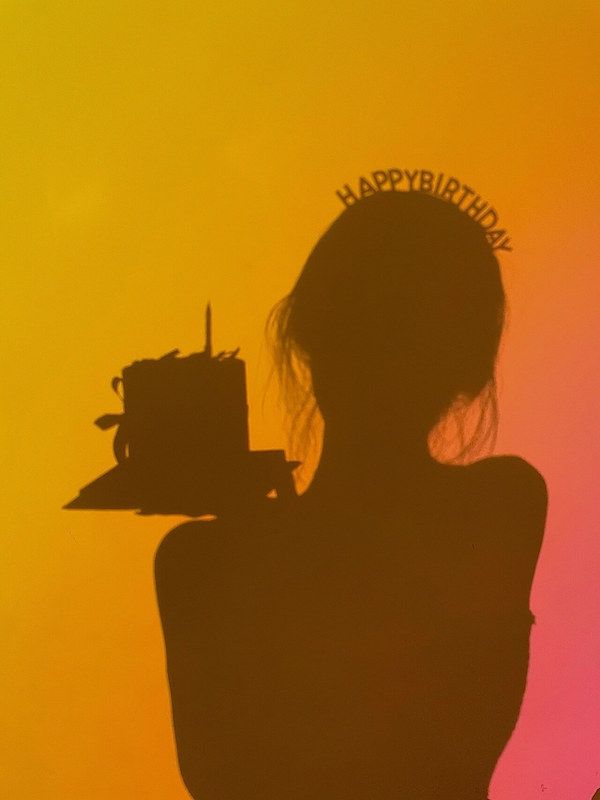 a silhouette of a girl holding a birthday cake with a "happy birthday" headband. the image is captured in front of a yelloworange wall with a slight pink hue. the light creates a beautiful silhouette effect, highlighting the celebratory mood of the scene.