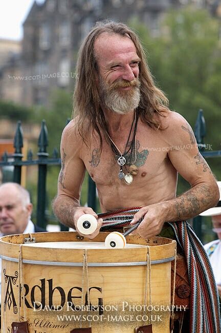 a man with long, dark, flowing hair, a beard and a long, patterned kilt plays a drum. he is shirtless, and has tattoos on his chest and arms. the drum is a traditional, wooden type with an ardbbeg inscription.