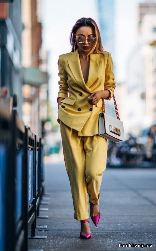 a stylish woman in a bright yellow pantsuit strolls down a city street. she's wearing fuchsia heels, sunglasses, and carries a white handbag.  the photo captures a trendy and confident urban fashion look. the background is blurred but shows city buildings and a hint of urban environment.