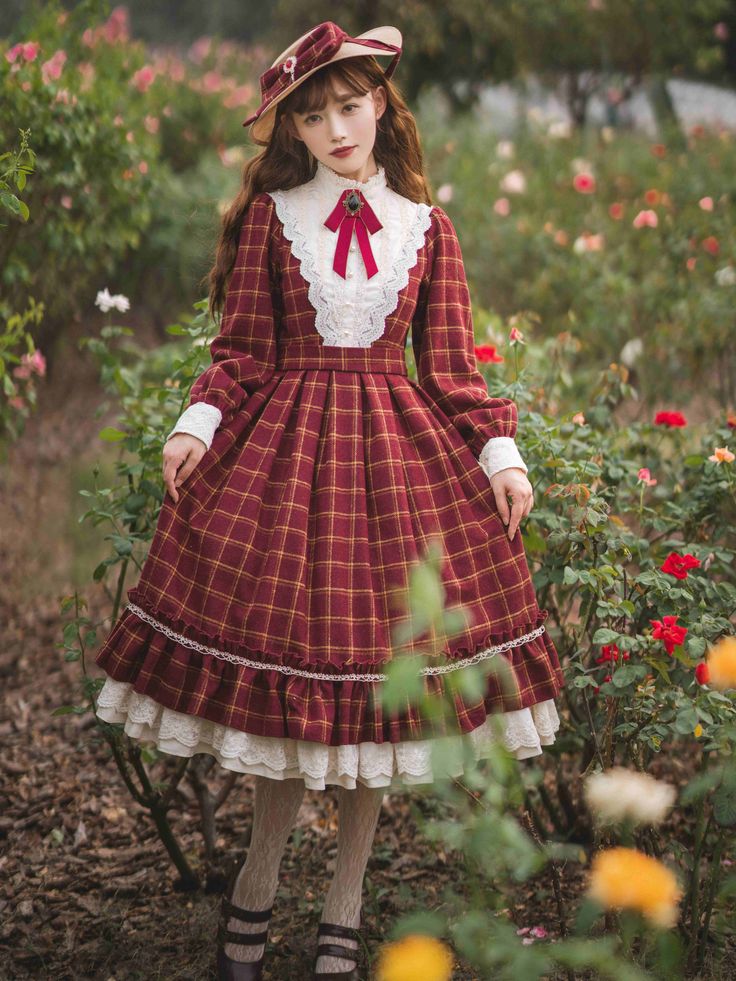 a woman in a vintageinspired red and yellow plaid dress stands in a garden. the dress has long sleeves, a lace collar and underskirt, and falls to her shins. she is wearing a matching hat and a red bow at her neck. the garden features red and pink roses in bloom. the photo is taken from eye level in bright daylight.