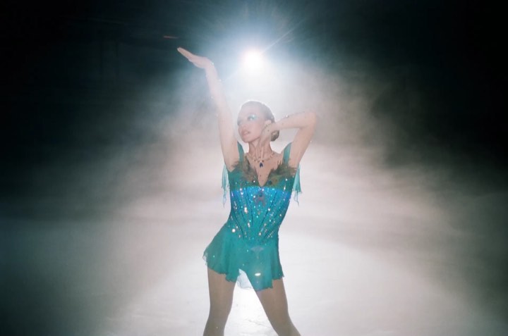 the image shows a female figure skater in a teal, sequined skating dress.  she's poised in a graceful pose on an ice rink, with a spotlight shining on her from above.  a light fog or smoke effect adds to the atmospheric and dramatic feel of the photograph. the skater appears to be in the midst of a performance, highlighting her elegance and athleticism.