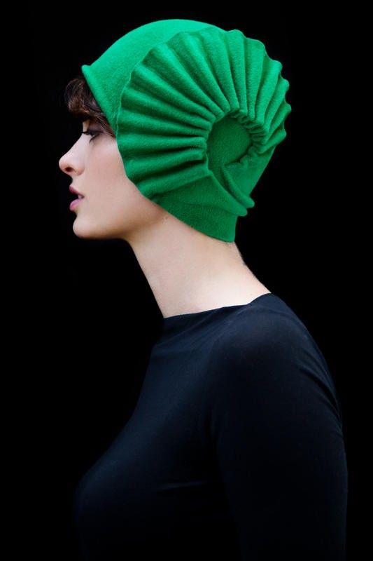a closeup portrait of a woman in a green hat, with a dark background. the hat is a soft, pillbox style with a scalloped edge. the woman is looking off to the side and her expression is serious.