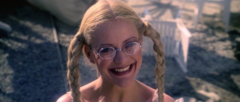 a blonde woman with pigtails and round glasses smiles brightly at the camera. she is outdoors and the image appears to be a closeup portrait.  she has on dark lipstick and is wearing a white lacy top.  the background is out of focus but appears to be natural and slightly industrial.