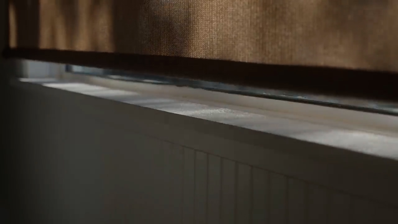 a closeup shot of a window with a brown roller blind partially drawn. the sunlight is streaming through the blind, casting shadows on the white window sill.