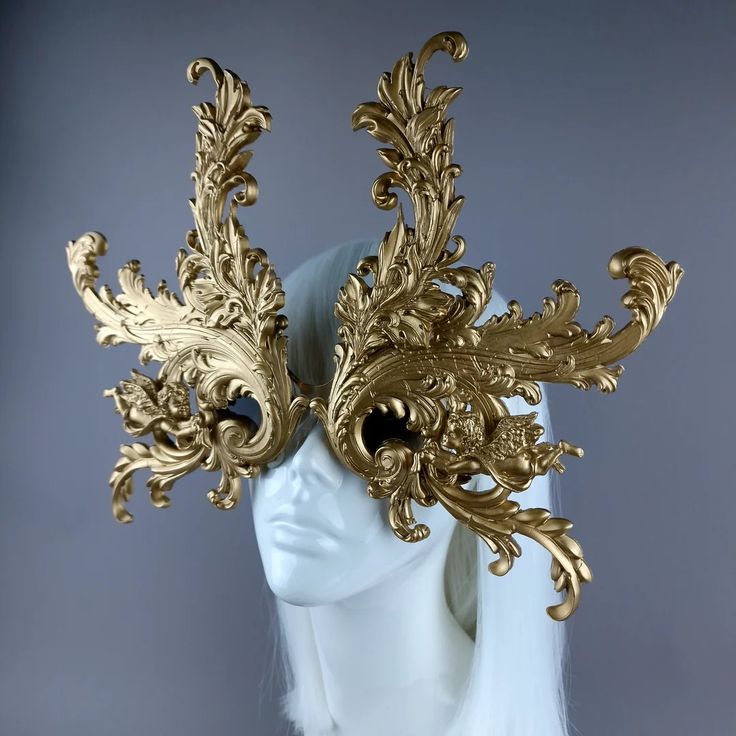 the image showcases a striking, goldcolored masquerade mask.  the mask features a highly intricate and ornate design reminiscent of baroque or rococo styles. it's large and dramatic, extending outwards in elaborate, swirling, leaflike patterns.  the mask is displayed on a white mannequin head with a white wig, providing a stark contrast that highlights the mask's details. the background is a simple, neutral gray, further emphasizing the mask as the focal point. the overall aesthetic is one of luxurious extravagance and theatrical flair.