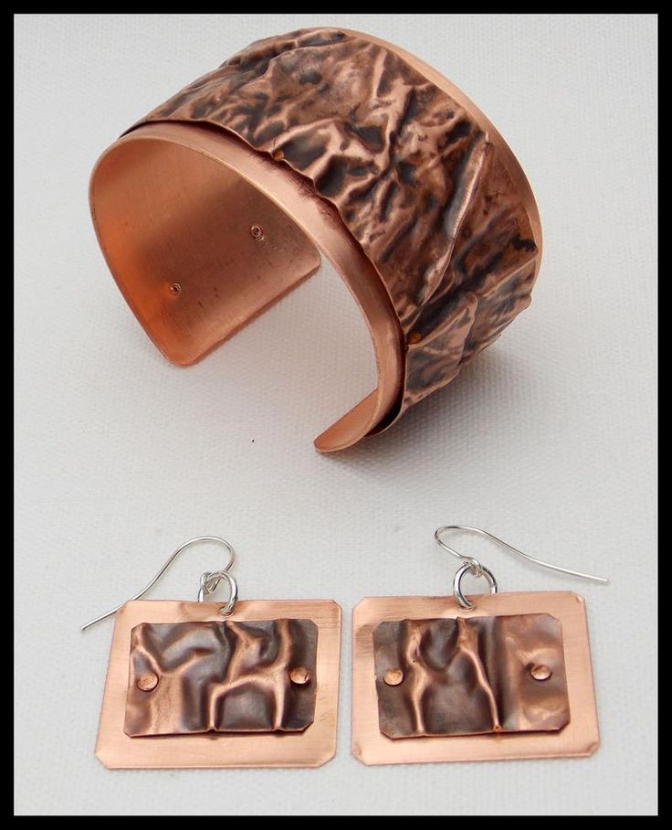 the image shows a set of handmade copper jewelry, including a wide cuff bracelet and a pair of square dangle earrings. the bracelet has a textured, hammered surface, and the earrings feature a smaller textured copper square riveted onto a larger smooth copper square, with silver ear wires.