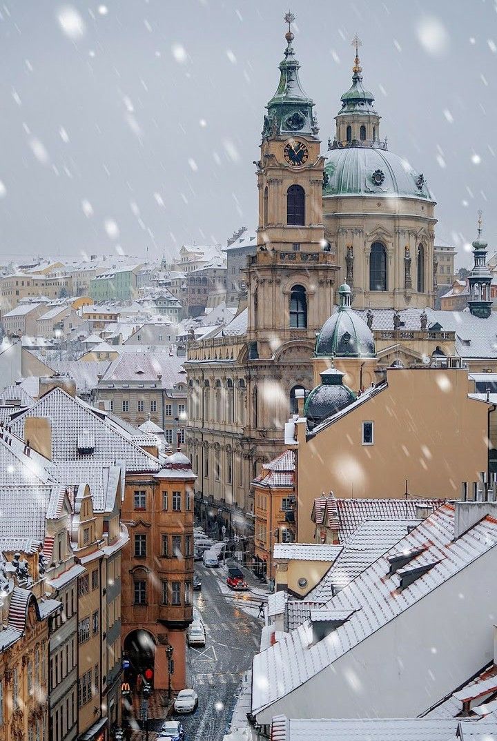 an aerial view captures a snowcovered prague cityscape, dominated by the st. nicholas church with its clock tower and dome. snow falls heavily over the roofs of buildings, which vary in color from tan to brown, and a street below with traffic lights and cars.