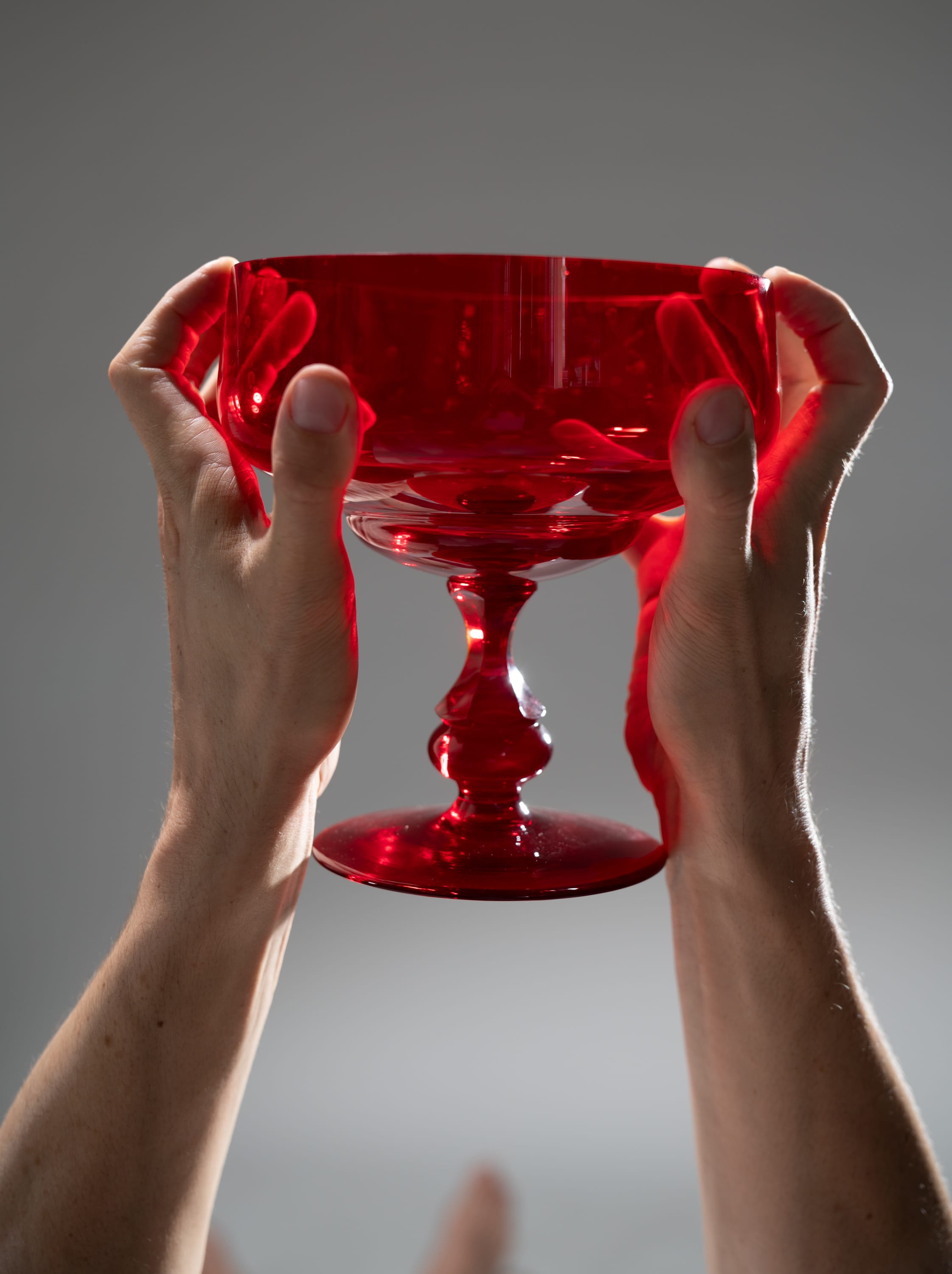 the image shows a pair of hands carefully holding a red glass chalice against a gray background. the glass appears to be empty and its intricate design is highlighted by the way the light shines through it. the low angle shot emphasizes the delicate balance and the vibrant red color of the glass.