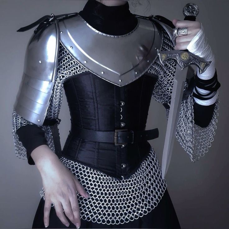 the image shows a woman dressed in a dark fantasy warrior costume. she's wearing a black corset, chainmail, silver pauldrons (shoulder armor), and a short black skirt. she holds a silver dagger and wears a black turtleneck. the overall aesthetic is dark, elegant, and powerful. the background is a simple gray. the image is welllit and captures the details of the costume clearly.