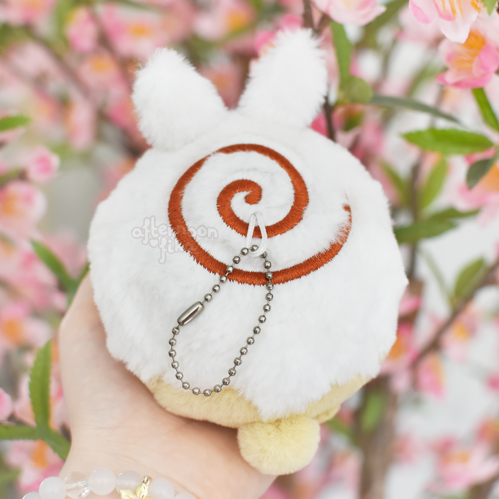 a closeup shot of a soft, white plush toy shaped like a rabbit with a cinnamon roll design on its back. the plush toy is attached to a silver keychain.  the hand holding the plush toy is visible, and the background is blurred but shows a blooming cherry blossom tree. the overall aesthetic is cute, pastel, and springlike.