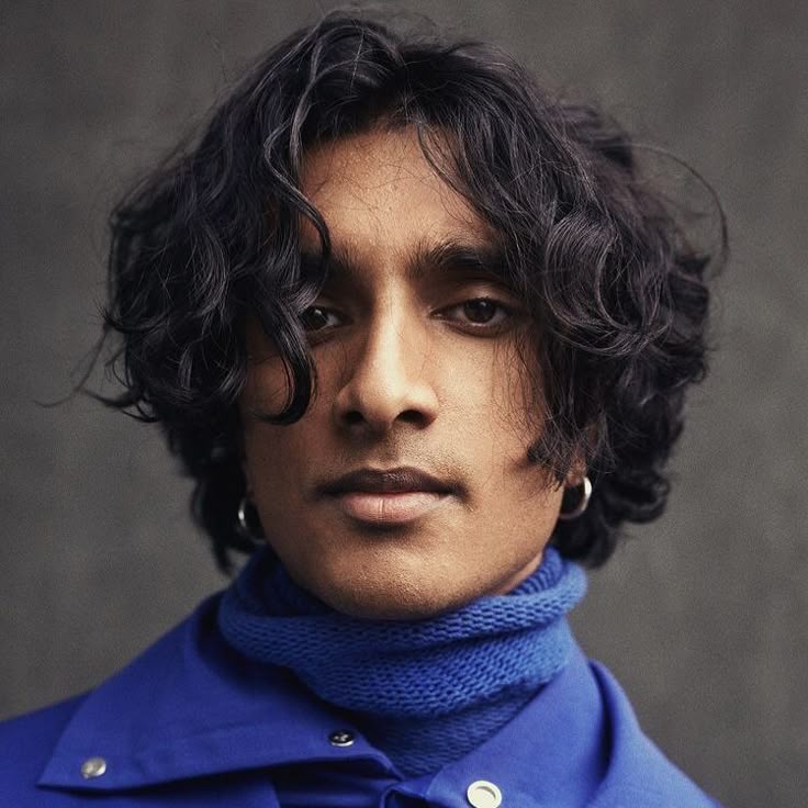 a portrait of a man with dark, curly hair wearing a blue turtleneck and jacket. he has an earring in his left ear and is looking directly at the camera. the background is a muted gray. the shot is a closeup, focusing on his face.