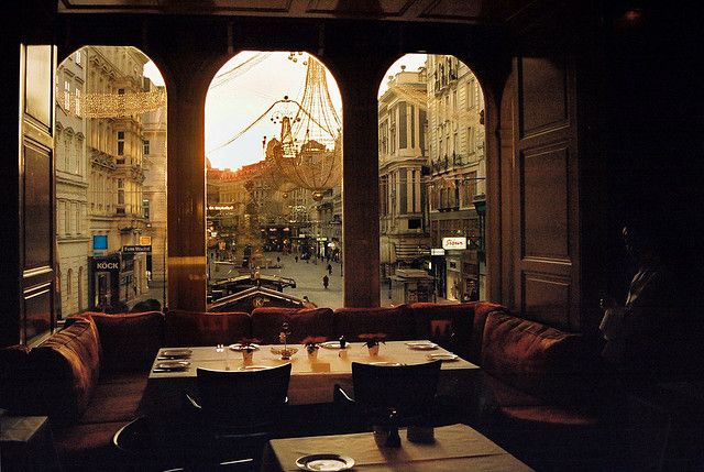 the image shows the interior of a sophisticated restaurant or cafe in vienna, austria.  large arched windows offer a stunning view of a city street at sunset.  the warm golden light of the setting sun illuminates the buildings and street scene outside, creating a dramatic contrast with the dark wood interior of the establishment.  inside, there's a comfortable boothstyle seating arrangement with a table set for a meal. the overall mood is cozy, romantic, and elegant, suggesting a highend dining experience.  holiday lights are visible in the street scene, hinting at a festive season. the photograph is well composed and captures a beautiful moment in time.