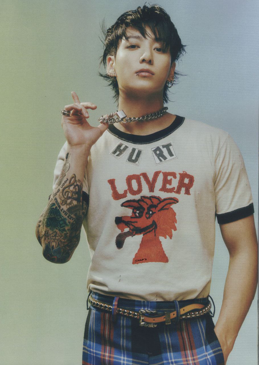 a portrait of a man with dark hair and tattoos, wearing a white tshirt with "hurt lover" printed on it, and blue plaid pants. he is wearing a chain necklace and has a lip piercing. he is holding a ring between his fingers.