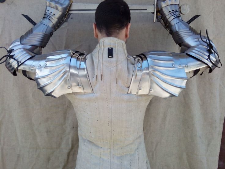 he image shows a person from behind wearing a suit of medieval plate armor. the armor includes pauldrons (shoulder guards), gauntlets (hand protection), and what appears to be portions of the arm guards and cuirass. the pauldrons are particularly prominent, featuring a fluted or scaled design. the person is also wearing a padded undershirt.  a sword is held behind their head in their hands. the background is a neutralcolored fabric backdrop.  the overall impression is one of authenticity and detail in the craftsmanship of the armor.