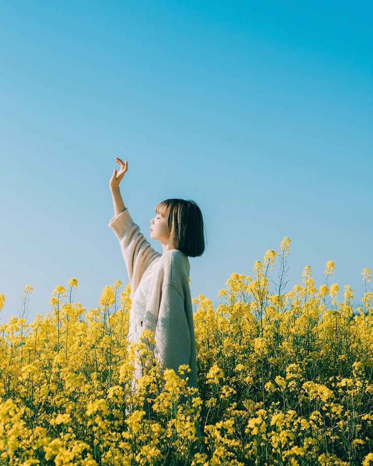 a young asian woman stands in a vibrant yellow field of rapeseed flowers, reaching her hand up towards the bright blue sky. she is wearing a light beige cardigan and has a calm and peaceful expression on her face. the sun is shining, and the overall mood of the image is one of serenity and happiness.