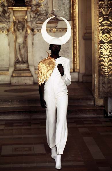 a fashion model with dark skin walks down a runway. she wears a white jumpsuit with gold embellishments on the shoulder and a large, white crescentshaped headdress. the background shows ornate architecture with stone pillars and arches.