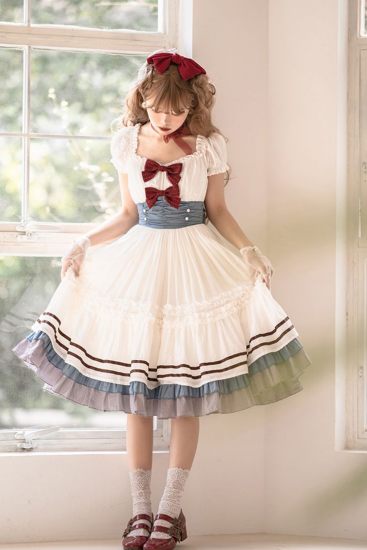 a young woman is wearing a white lolita fashion dress. the dress has a square neckline, puffed sleeves, a blue waistband, and a layered skirt with brown stripes. she is also wearing a red bow in her hair, white socks, and red mary jane shoes. she is standing in front of a window, and the lighting is soft and natural.