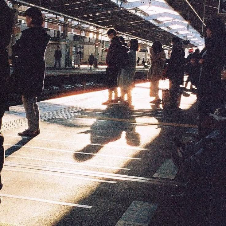 the image is a long shot of a train station platform bathed in sunlight streaming through the high, metal roof.  several people, mostly dressed in winter coats, are waiting on the platform, their long shadows cast dramatically by the low sun. the focus is on the individuals waiting and their shadows, creating a somewhat melancholic yet peaceful atmosphere. the overall aesthetic suggests a scene in a japanese train station. the background shows a partially visible train track and furtheroff figures. the scene shows a moment of quiet anticipation before a train arrives.