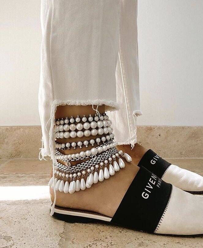 a close up of a woman's ankle wearing several white pearl and silver chain anklets.  she is wearing white jeans that are ripped at the ankles, white and black sandals that say "givenchy" on the side, and has the bottom half of her leg and foot showing.  the picture is focused on the ankles and the fashion choices she has made.