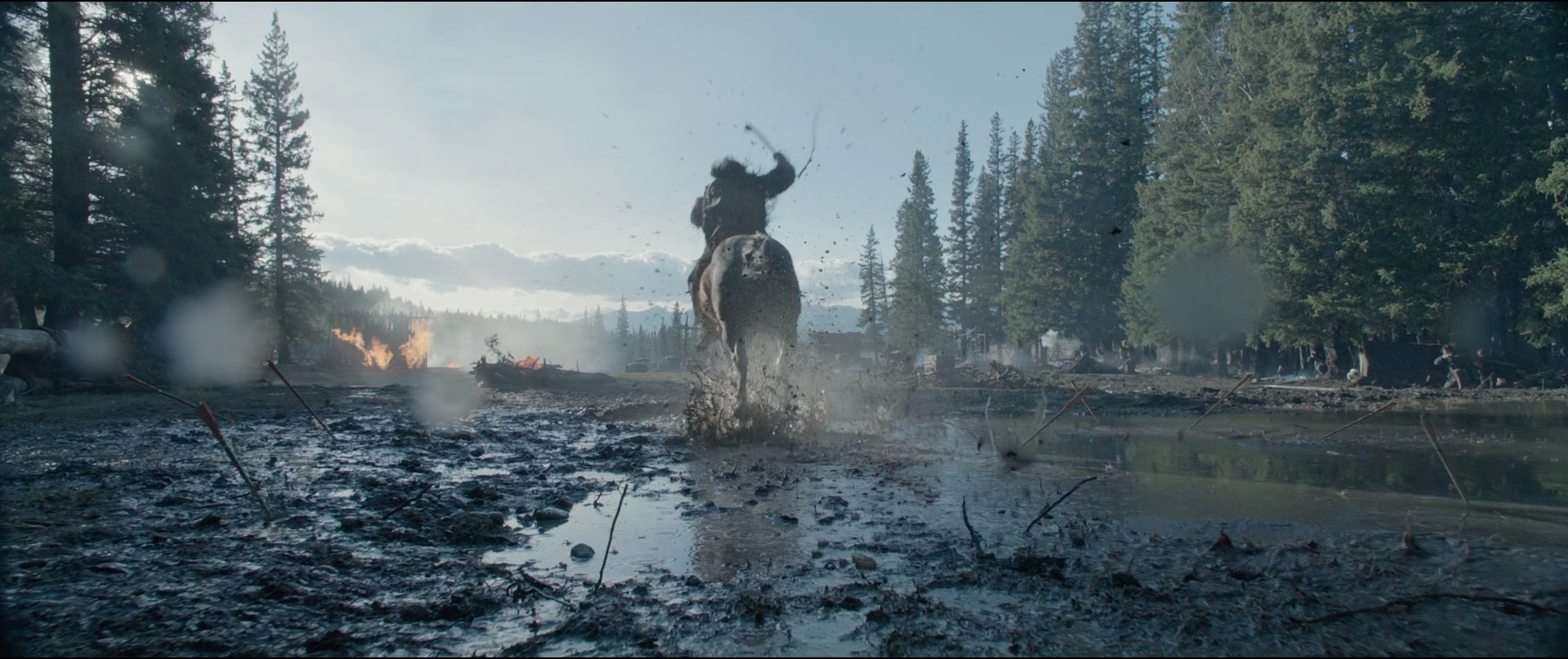 a man riding a horse through the mud with a weapon raised. there are trees and a fire in the background. the image is taken from a low angle.