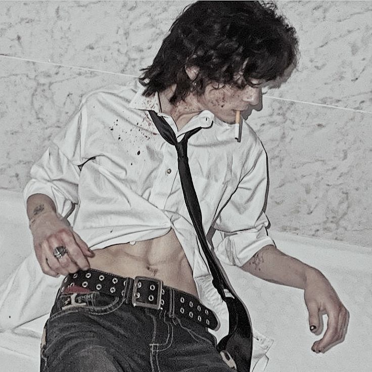 a man is posing for a fashion shoot, wearing a white shirt, black tie, black jeans, and a belt. he is holding a cigarette in his hand, and his hair is dark and styled. the background is white.