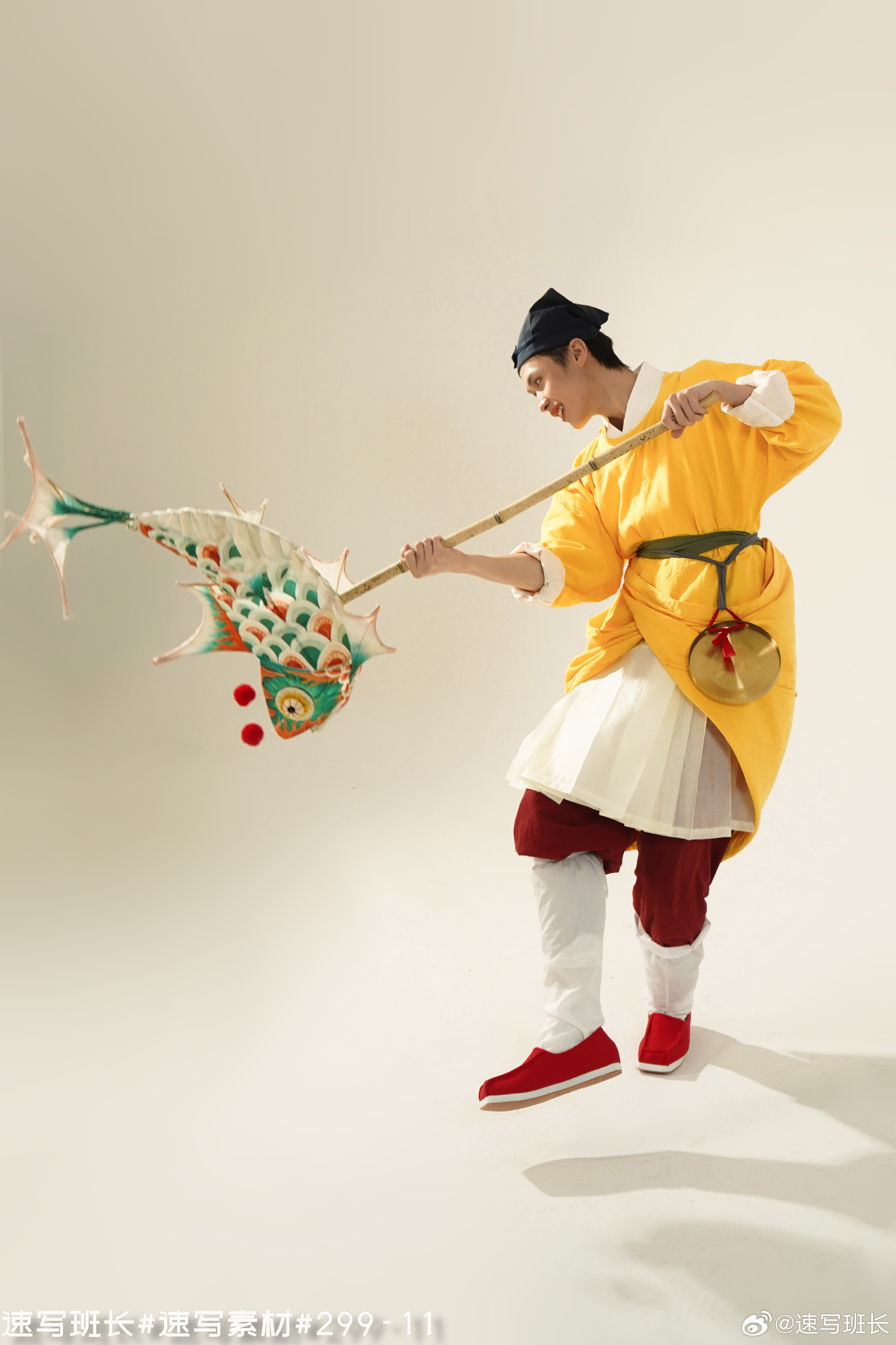 a young man in a traditional yellow and white chinese costume is dynamically posed against a white backdrop. he holds a long bamboo pole from which a colorful, fishshaped kite dangles. he wears red shoes and a black cap, and a golden cymbal hangs from his waist. the image evokes a sense of joyous celebration and cultural performance, likely relating to chinese new year or spring festival.