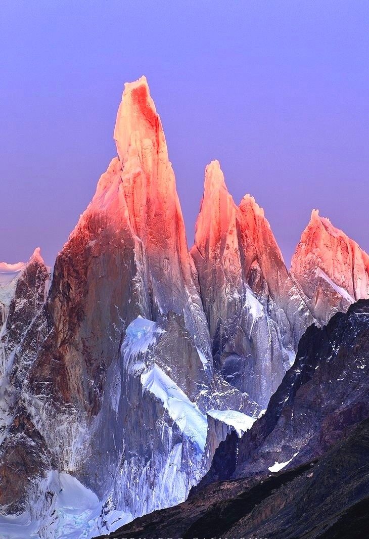 the image shows a stunning mountain landscape at sunrise. several jagged, rocky mountain peaks dominate the scene, their tops illuminated with a bright pink hue, likely from the rising sun. the lower portions of the mountains are covered in snow and ice, adding to the rugged beauty of the landscape. the sky is a soft, pale purple, further enhancing the colors of the scene.