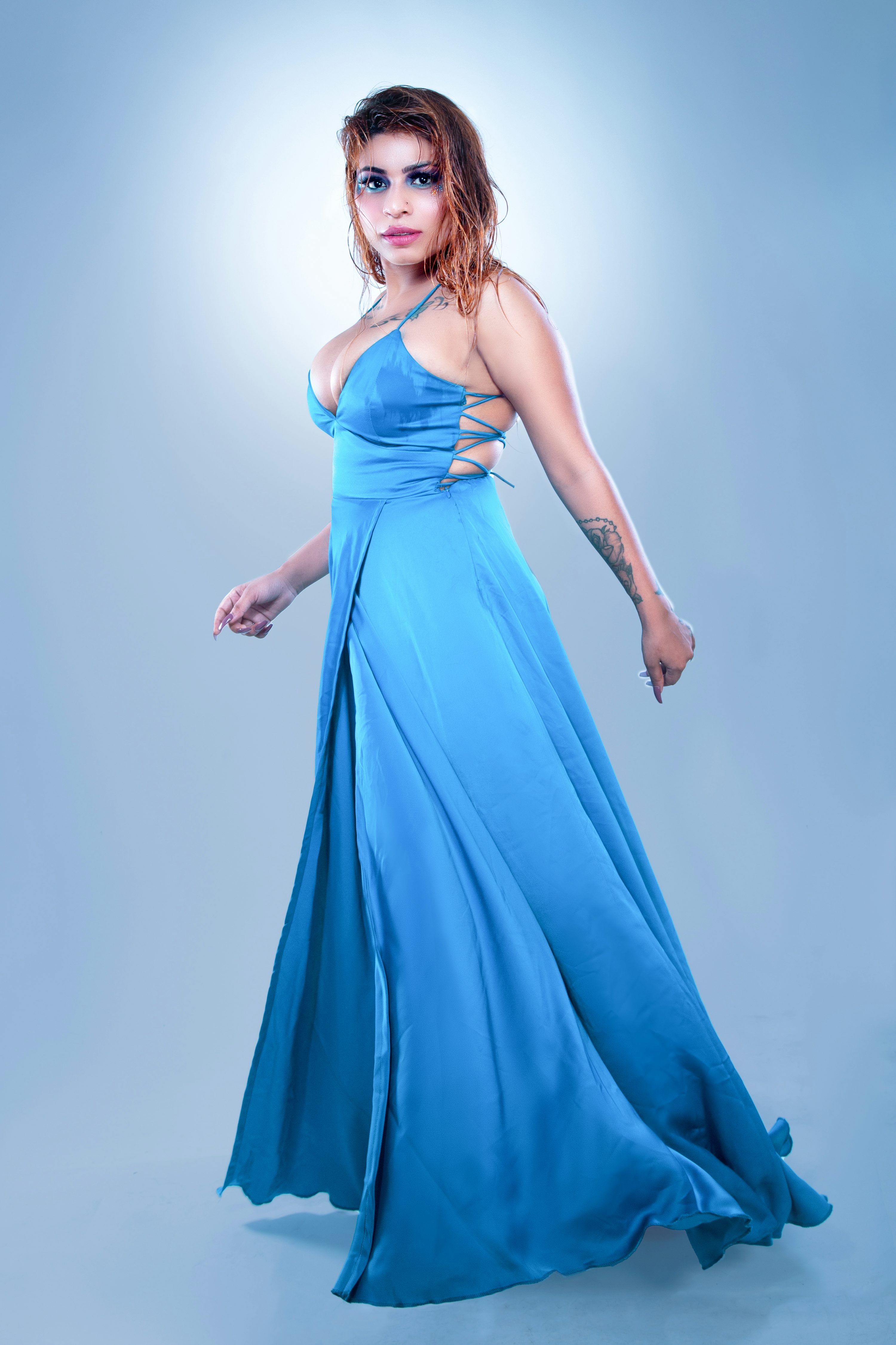 a fulllength studio shot of a woman wearing a flowing teal maxi dress. the dress is sleeveless with a low vneck and features thin straps that crisscross in the back. the woman has long wet brown hair and makeup that enhances her features. she is posing with confidence and grace, her dress flowing elegantly around her. the background is a light blue gradient.