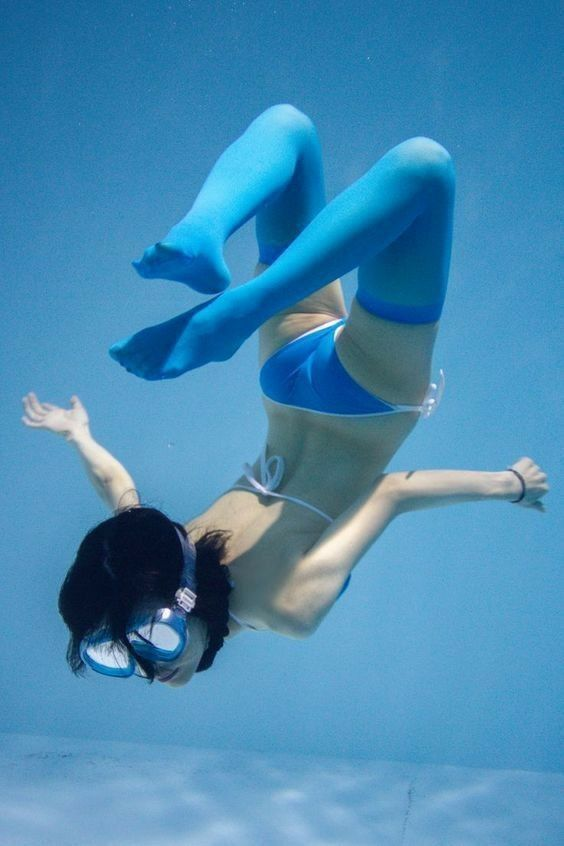 a young woman is captured in a dynamic underwater photograph. she's submerged in a bright blue pool, wearing a blue and white bikini and blue swim stockings.  her pose is playful and energetic, with her limbs extended as if caught midmovement. she wears swim goggles, her dark hair fanning out around her head.  the lighting is artificial and bright, emphasizing the clarity of the water. the overall impression is one of joy and freedom.